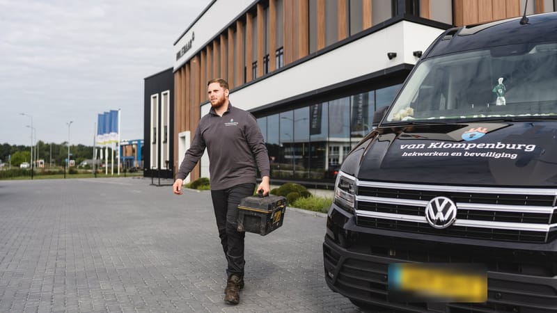 Monteur loopt met zijn gereedschap naar een bedrijfspand, met de bedrijfsbus op de voorgrond